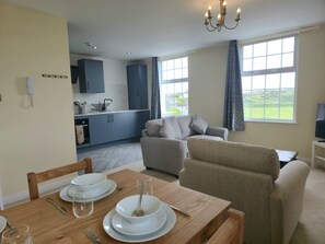 A dining area with a table set for two, a sofa, and a kitchen in the background. - 1 Bedroom + Sofa bed in centre of Trearddur Bay - Pass the Keys (Bae Trearddur)