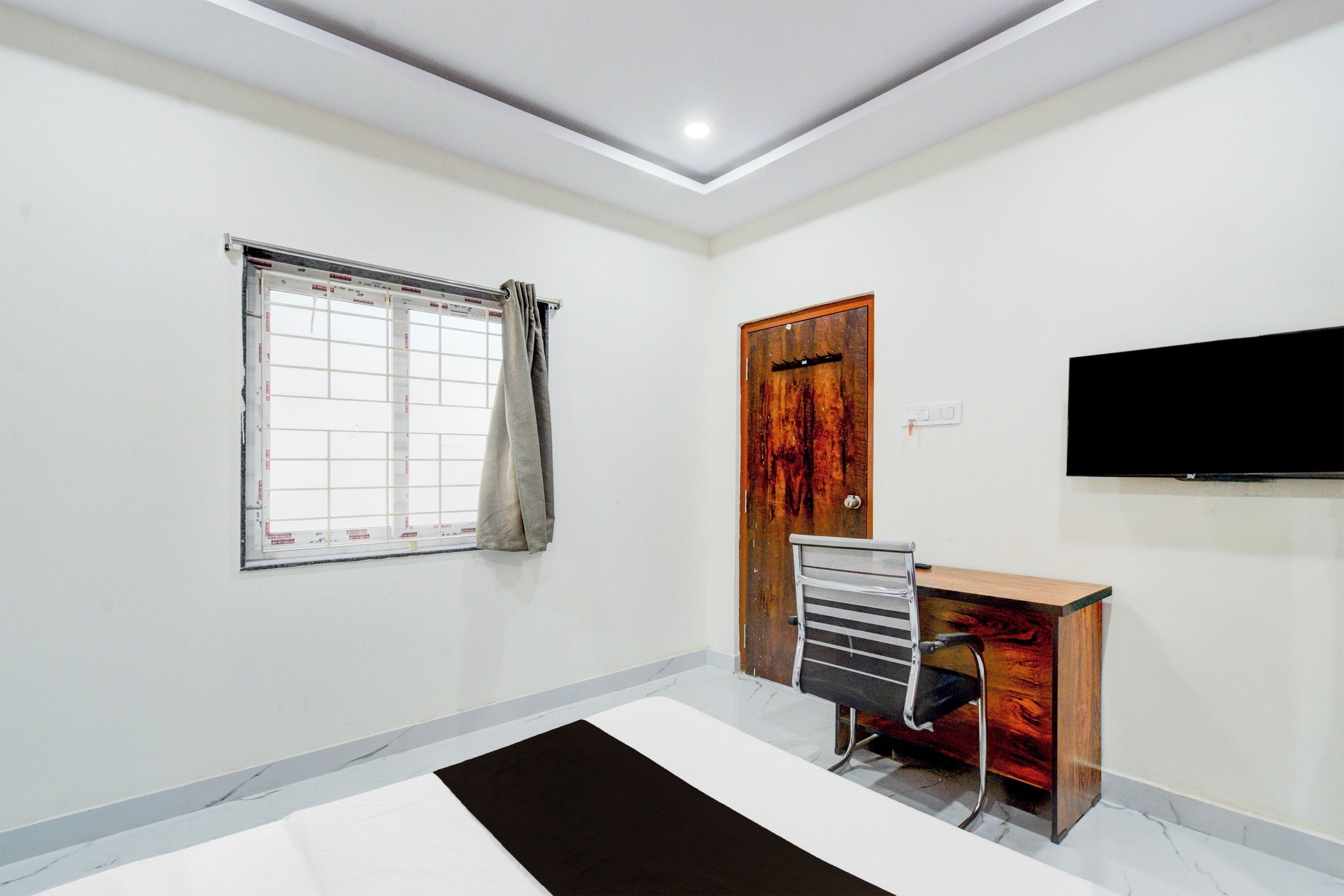 Quarto com cama, escrivaninha, cadeira, janela com cortinas, porta e TV de tela plana fixada na parede.