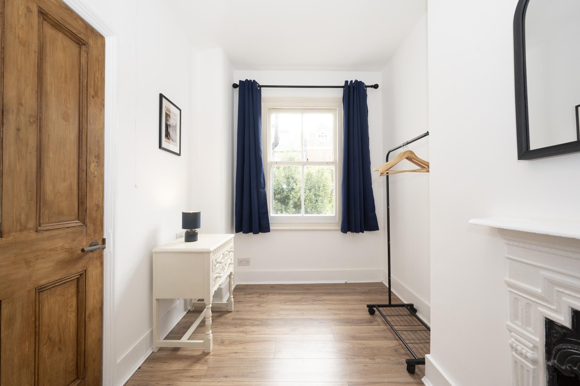 A hallway with a wooden door, a white dresser, a window with blue curtains, and a coat rack.