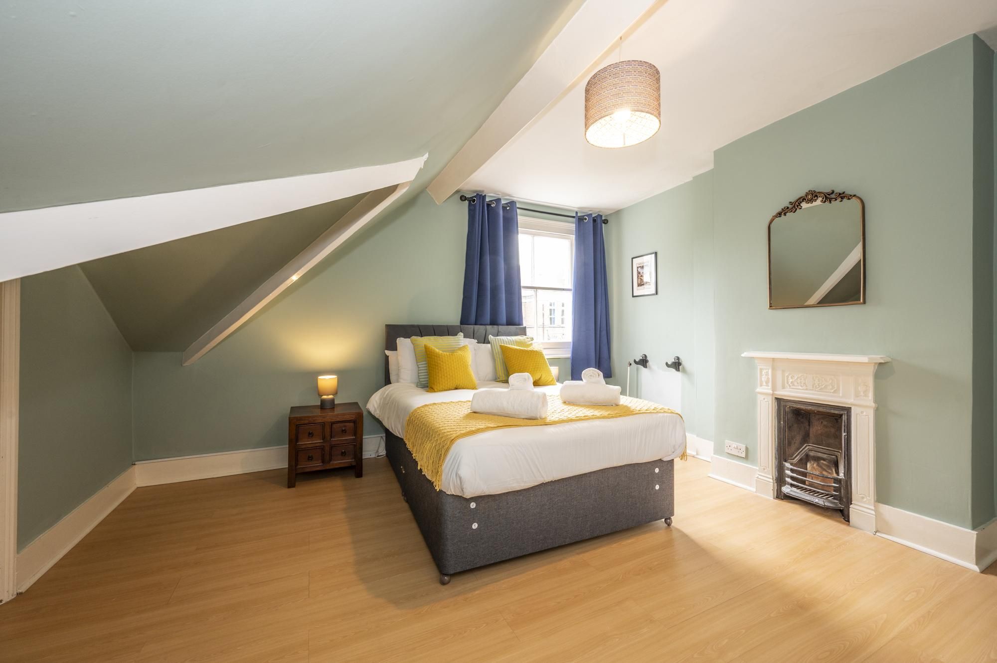 A bedroom with a bed, a nightstand, a mirror, a fireplace, and a window with curtains.