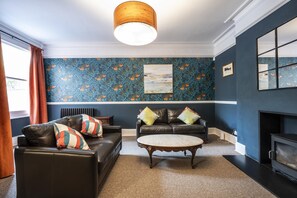 Ein Zimmer mit dunkelblauer Wand, einem gemusterten Wandteppich, einem Kamin, einem Ledersofa, einem Couchtisch und einer hängenden Leuchte.