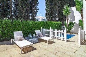 Uma área de estar externa com duas poltronas e uma mesa pequena, um cercado branco e uma piscina ao fundo.
