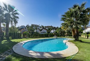 A round swimming pool with a clear blue water surface, surrounded by a well-maintained lawn and palm trees. - AR23 - Holiday flat, Puerto Banus by Roomservices (Marbella)
