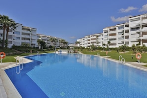 A swimming pool with a clear blue water surface, surrounded by a white building with balconies and palm trees. - Beachside Apartment with Pool, Puerto Banus- PR20 (Marbella)