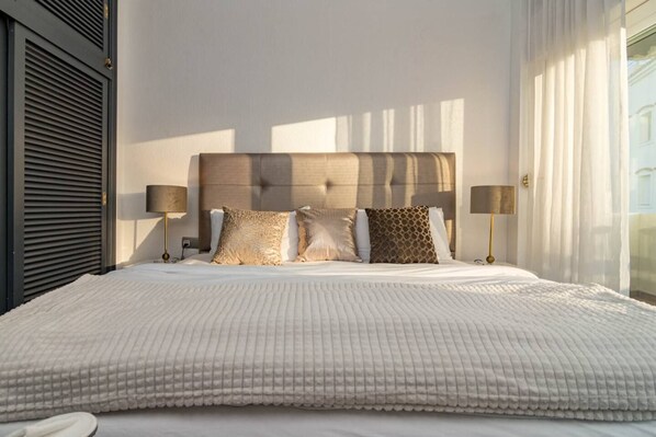 Un grand lit avec une couette blanche à motifs, une tête de lit en bois et deux lampes de chevet.