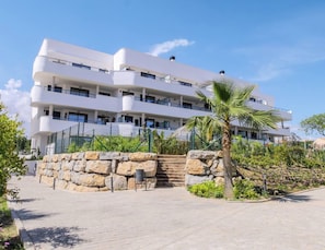 Un immeuble à plusieurs étages doté d’une architecture moderne, avec des balcons et un espace paysager agrémenté d’un jardin et d’un palmier.