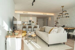 A modern living room with a sofa, coffee table, and chairs, a dining area with a table and chairs, and a kitchen in the background. - Sea-View 2BR, Steps to Beach & Old Town - EB4A (Estepona)