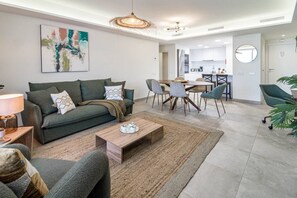 A modern living room with a sofa, coffee table, and chairs. There is a dining area with a table and chairs. The room has a neutral color palette and a large painting on the wall. - INf31J - Stunning holiday home next to beach (Estepona)