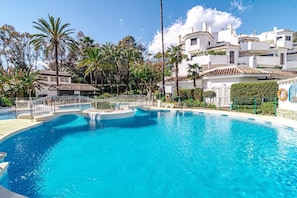 A swimming pool with a white deck, surrounded by palm trees and white buildings with red-tiled roofs. - GBH - Casa Golden beach by Roomservices (Marbella)