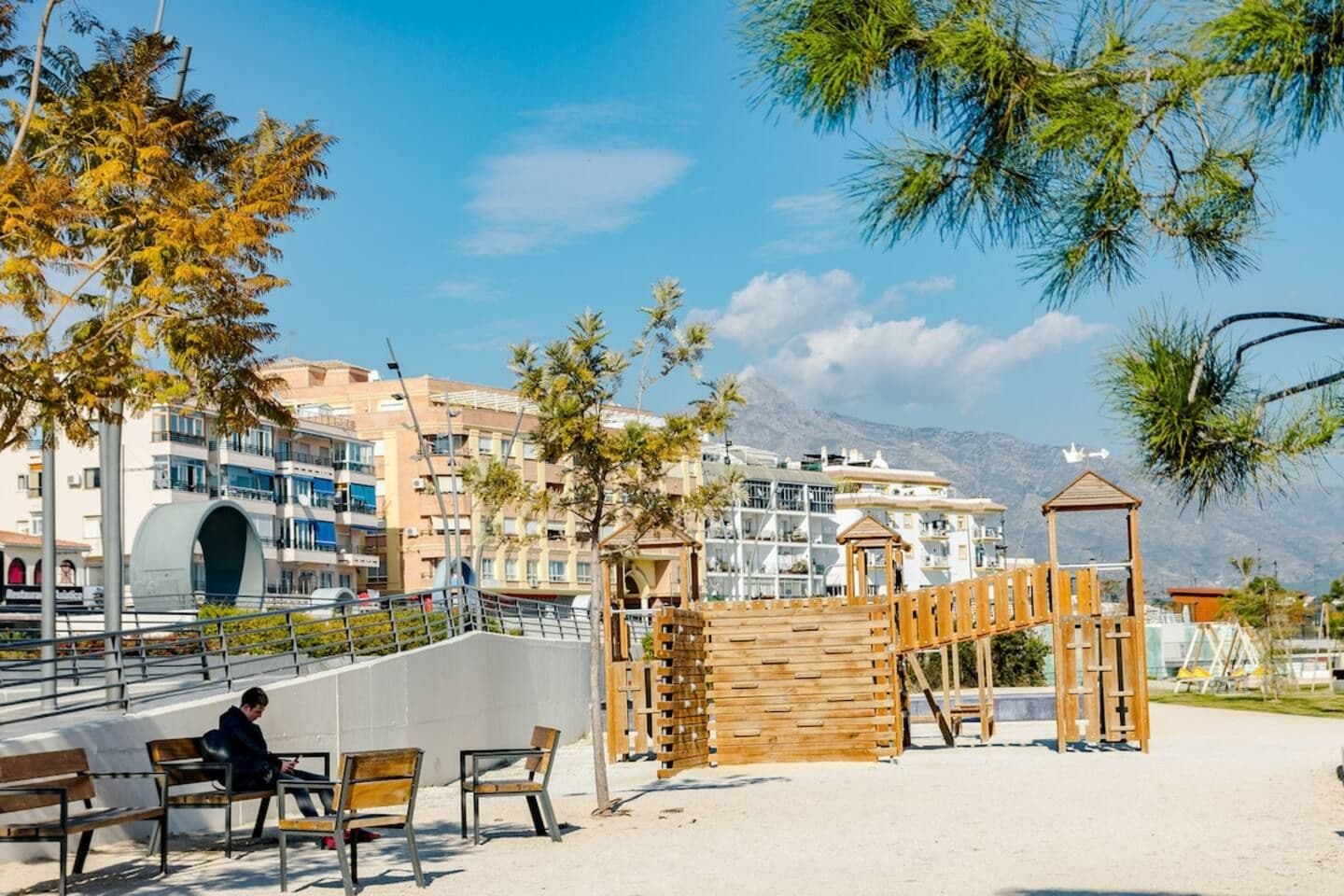 A beachside park with a wooden play structure, benches, and a view of residential buildings in the background.