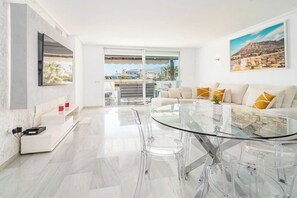 Una sala de estar moderna con una mesa de comedor de vidrio, sofá blanco y un amplio ventanal con vistas a edificios y montañas.