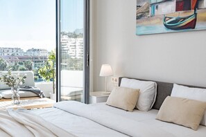 A hotel room with a large bed, a bedside table with a lamp, a view of the city through a large window, and a painting on the wall. - Modern Nueva Andalucia Apt, Pool & Gym - JG (Marbella)