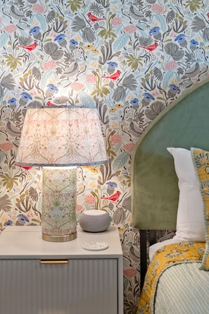 Ein ordentlich bezogenes Bett mit einem grünen Kopfteil, ein Nachttisch mit Lampe und eine Wand mit Blumenmuster.