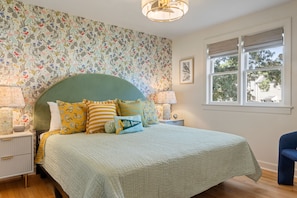 A bedroom with a floral wallpaper, a green headboard, a bed with striped bedding, two bedside lamps, and a window with a view of trees. - Lively Oak 175 Steps to The Village  Near NCSU (Raleigh)