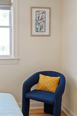 Ein Zimmer mit blauem Sessel, gelbem Kissen, einem gerahmten Blumenbild an der Wand und einem Fenster mit Jalousien.