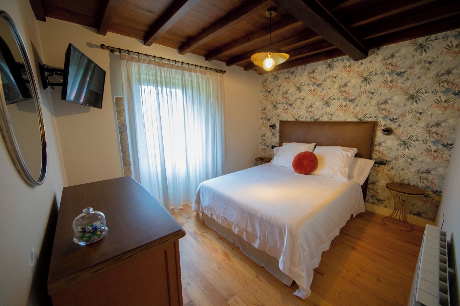 Un dormitorio con cama, mesita de noche, espejo, ventana con cortinas, pared con papel tapiz floral y techo de madera.