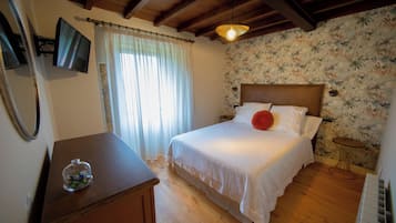 Un dormitorio con cama, mesita de noche, espejo, ventana con cortinas, pared con papel tapiz floral y techo de madera.