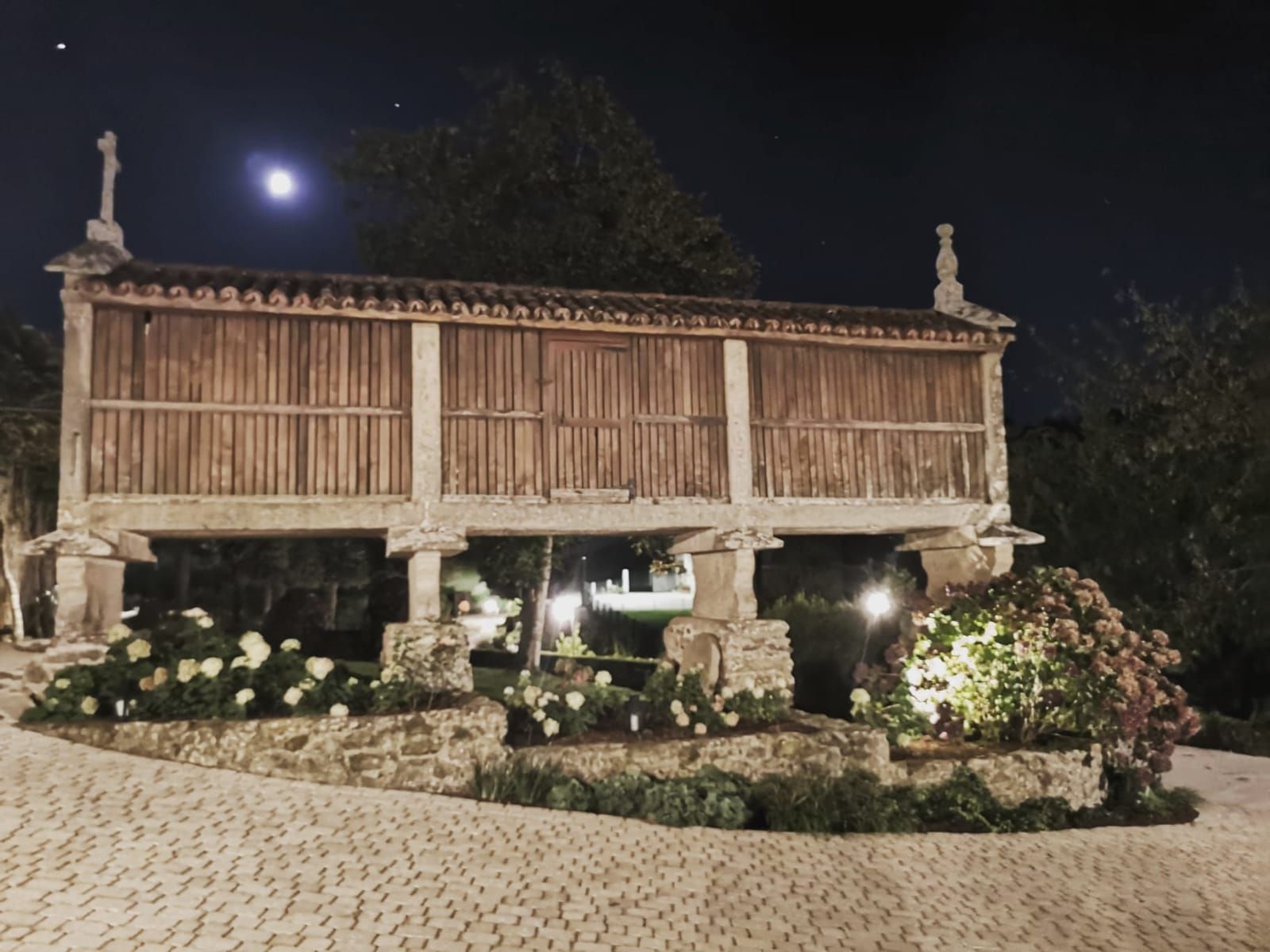 Ein Steinbauwerk mit Holzdach, umgeben von einem Garten mit weißen Blüten und viel Grün.