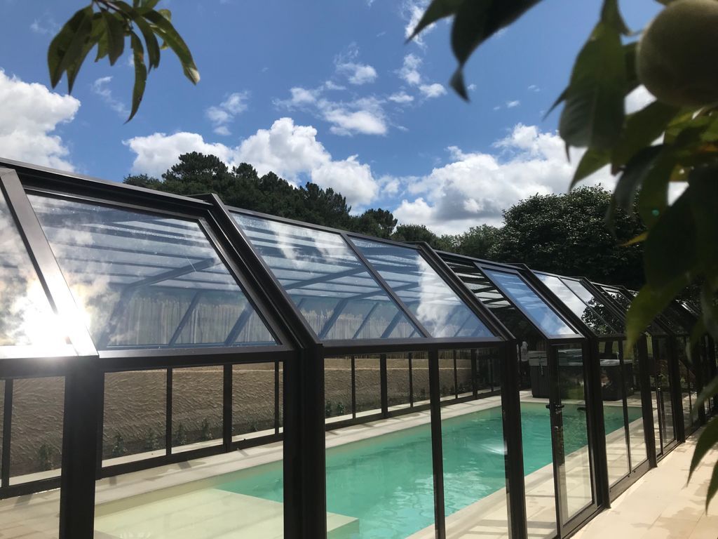Una piscina cubierta por un cerramiento de vidrio, bajo un cielo azul despejado con nubes.