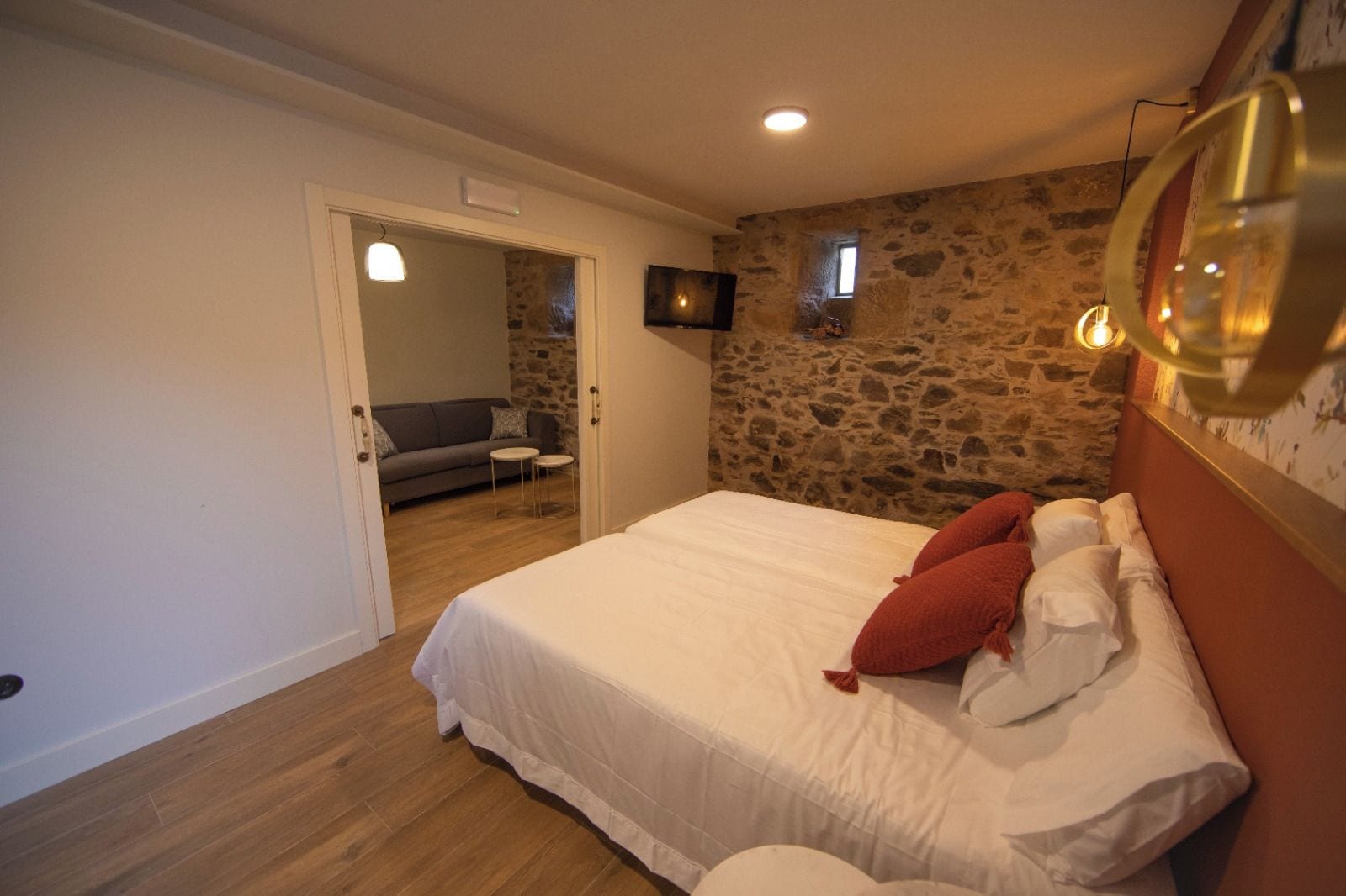 Un dormitorio con pared de piedra, cama con ropa blanca y almohadas rojas, un televisor pequeño en la pared y suelo de madera.