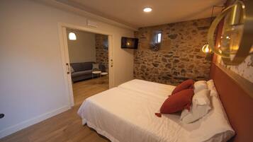 Un dormitorio con pared de piedra, cama con ropa blanca y almohadas rojas, un televisor pequeño en la pared y suelo de madera.