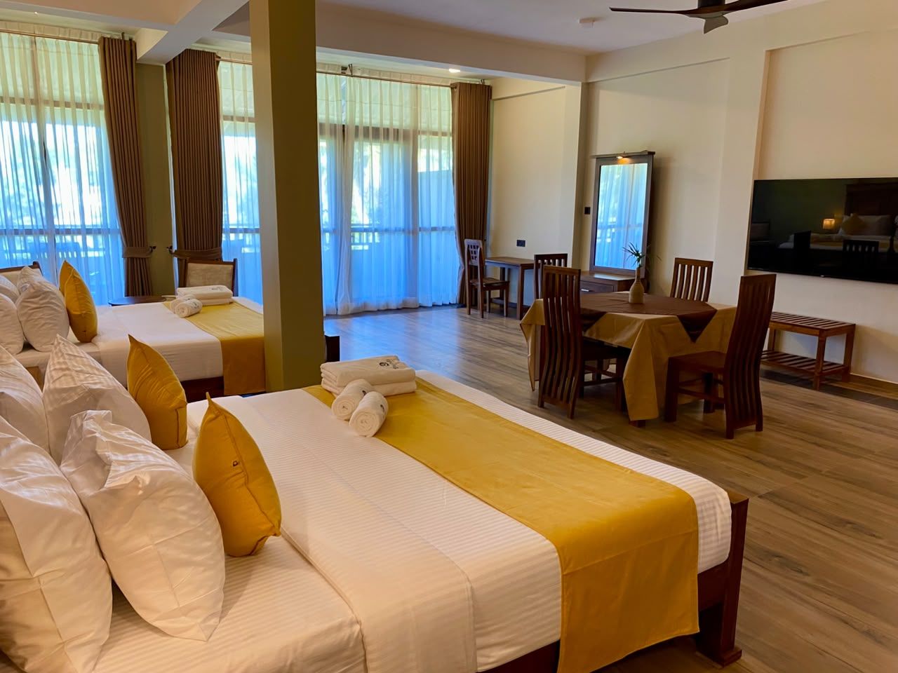 A hotel room with two beds, a dining table, chairs, a TV, and large windows with curtains.
