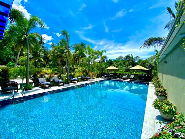 A swimming pool with lounge chairs and umbrellas, surrounded by palm trees and greenery.