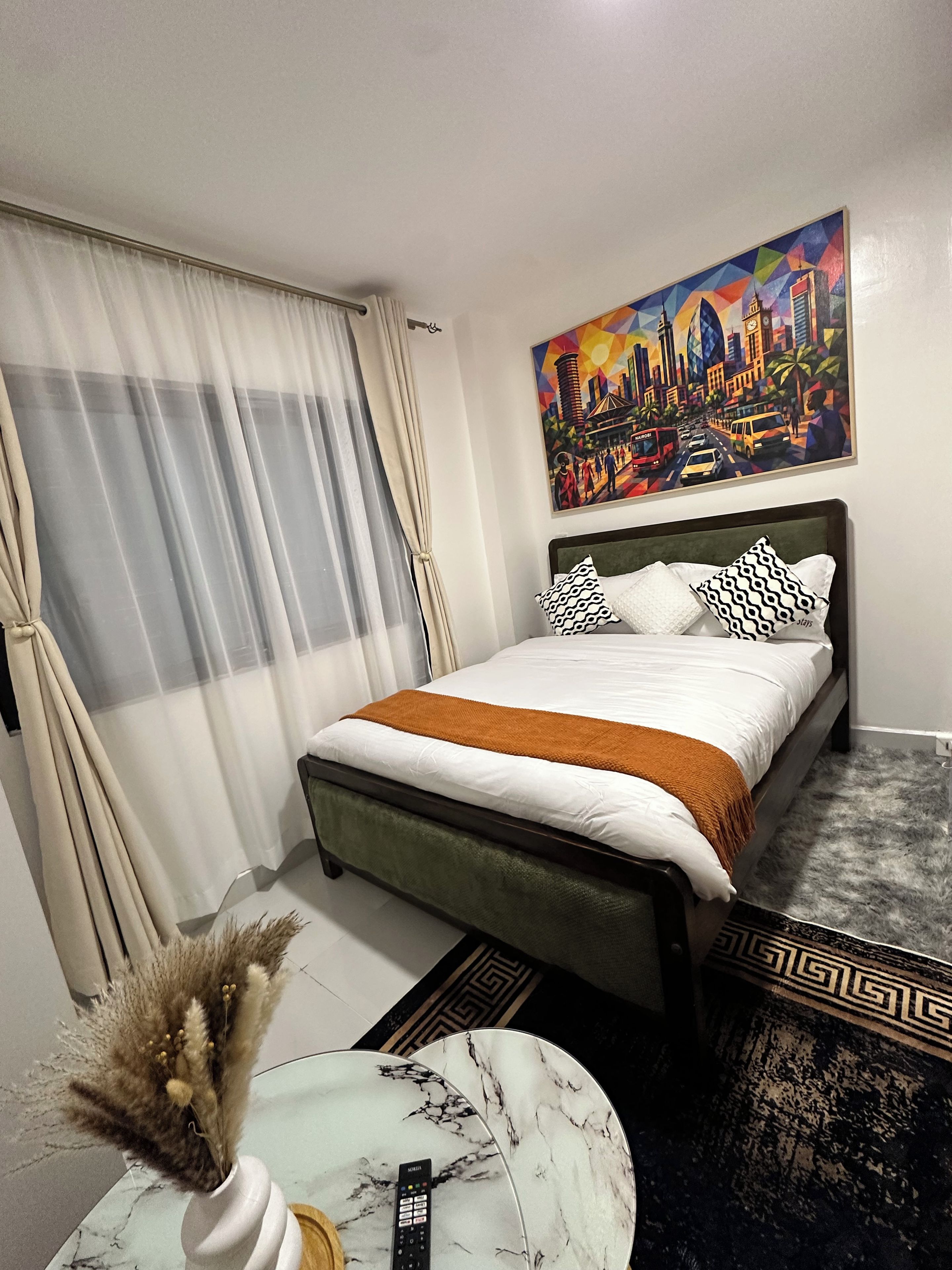 A hotel room with a bed, a painting, a remote control, and a decorative vase.