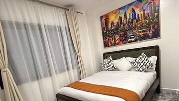 A hotel room with a bed, a painting, a remote control, and a decorative vase.