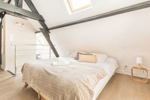 Un dormitorio con techo inclinado, una cama con colcha color beige, una lámpara de noche y piso de madera.