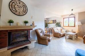 A living room with a fireplace, leather armchairs, a sofa, a dining area, and a large wall clock. - Le Repaire Paisible - Charmante maison pour 12 (Charny-Orée-de-Puisaye)