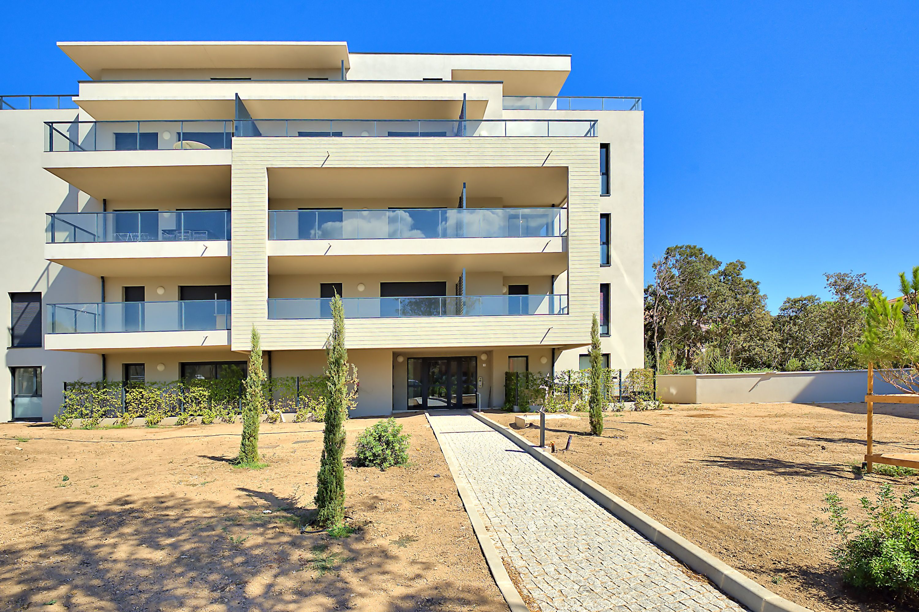 A multi-story residential building with balconies, a paved entrance area, and a landscaped garden.