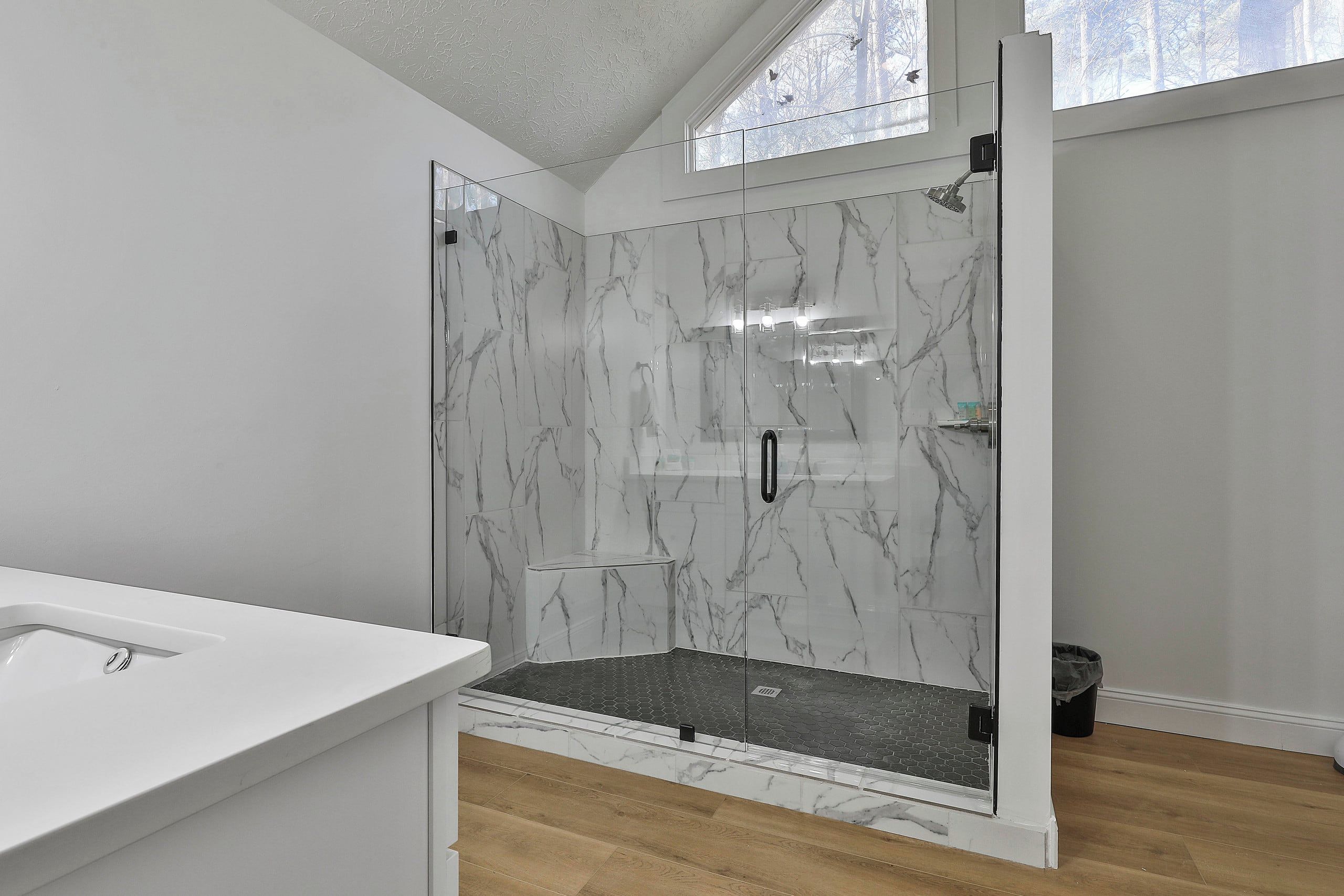 A modern bathroom with a glass-enclosed shower, marble walls, and a wooden floor.