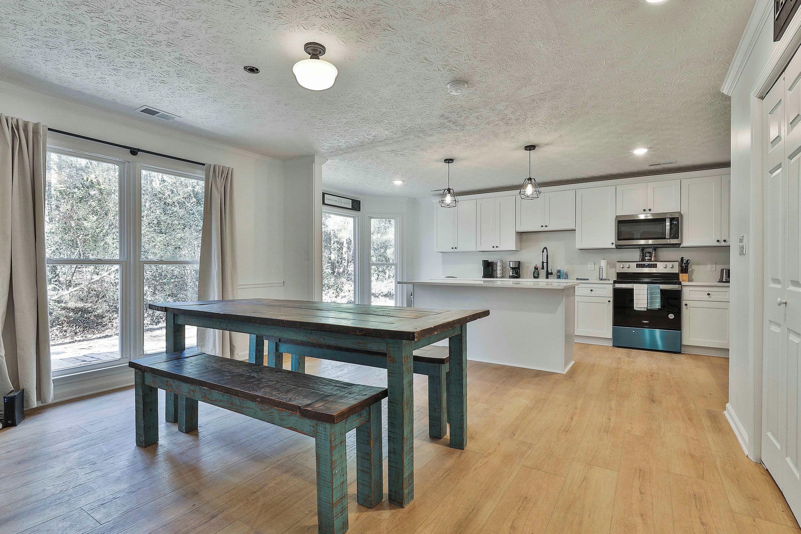 Una cocina moderna con armarios blancos, horno empotrado y un comedor con mesa y banco de madera.