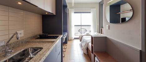 A compact hotel room with a kitchenette, a bed, a small balcony, and a round mirror.