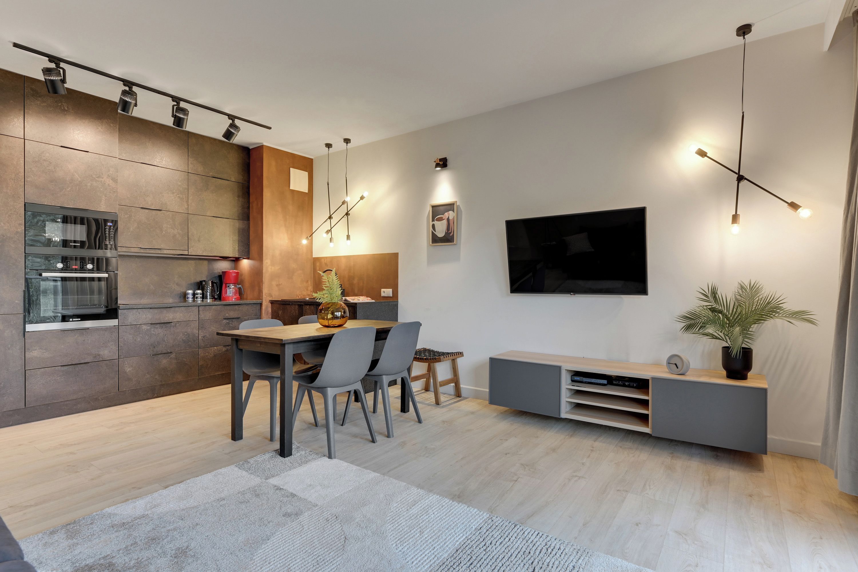 Una cocina moderna con electrodomésticos integrados, una zona de comedor con mesa y sillas de madera, un televisor de pantalla plana en la pared y un mueble de entretenimiento minimalista.
