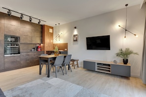 Una cocina moderna con electrodomésticos integrados, una zona de comedor con mesa y sillas de madera, un televisor de pantalla plana en la pared y un mueble de entretenimiento minimalista.