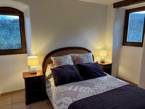 Een slaapkamer met een bed, twee bedlampen en uitzicht op bomen door een raam.