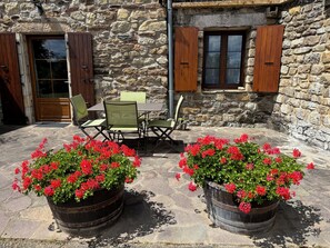 Een stenen gebouw met een patio, voorzien van een tafel en stoelen, en twee grote bloempotten met rode bloemen.