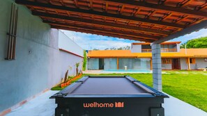 A pool table with 'welhome' branding, a covered outdoor area, and a building with orange roofing in the background. - Sitio em Mairipora com Piscina e Churrasqueira (Mairiporã)