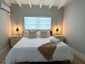 A neatly made bed with white bedding and a brown throw blanket, two bedside tables with lamps, a window with blinds, and an air conditioning unit on the wall. - Studio on the Barrier Island. Walk to the Beach (Fort Lauderdale)