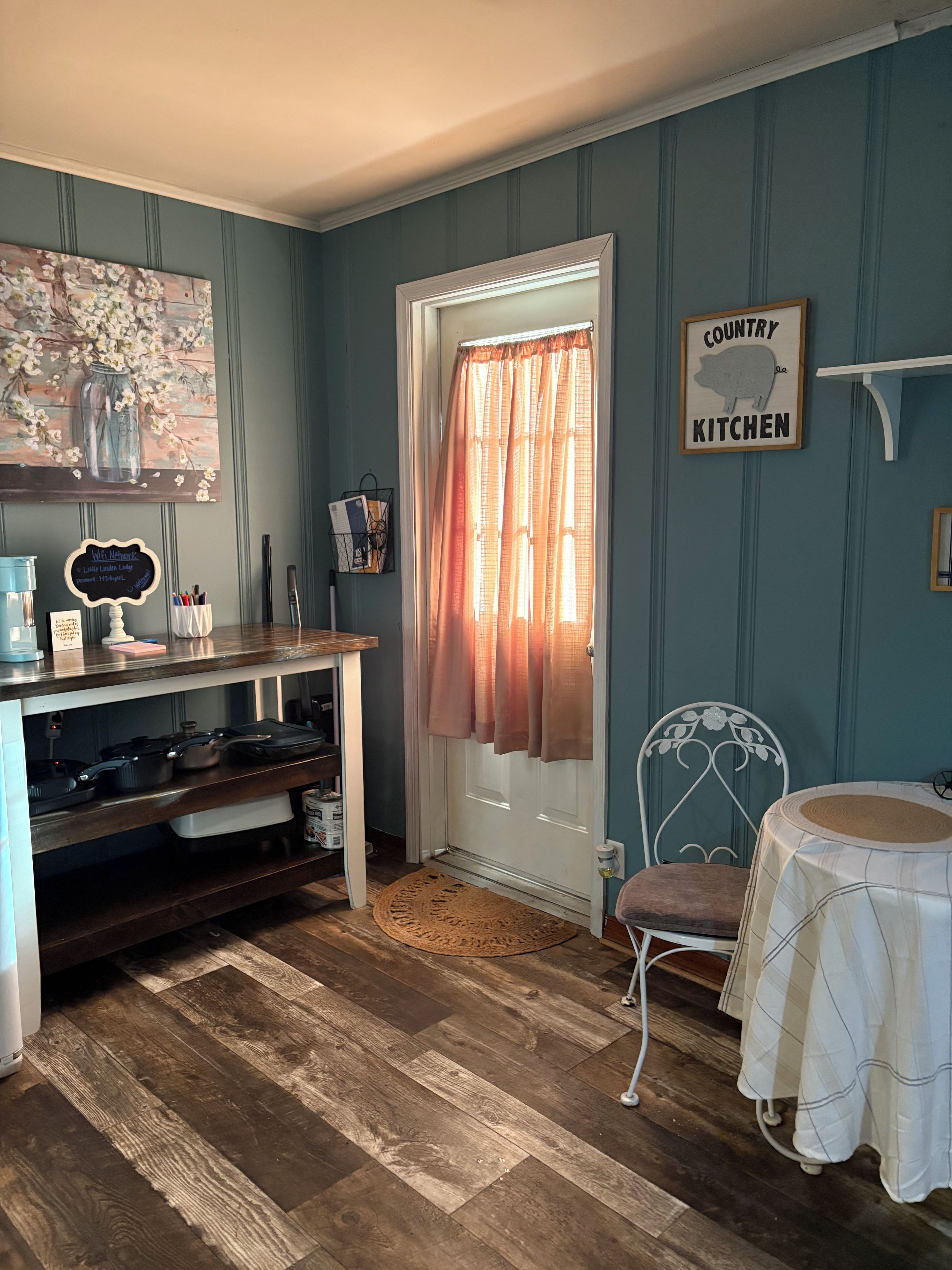 Una stanza con pavimento in legno, una porta bianca con tenda, una sedia in metallo, un tavolino con tovaglia bianca e un muro con il cartello 'Country Kitchen'.