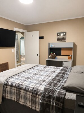 A bedroom with a bed, a bedside table with a lamp, a television mounted on the wall, and a door leading to another room. - Little Linden Lodge: 2-bedroom house in charming McKenzie (McKenzie)