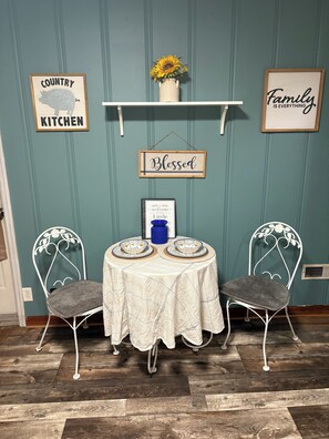 A dining area with a table set for two, two chairs, a wall-mounted shelf with a sunflower, and three framed signs. - Little Linden Lodge: 2-bedroom house in charming McKenzie (McKenzie)
