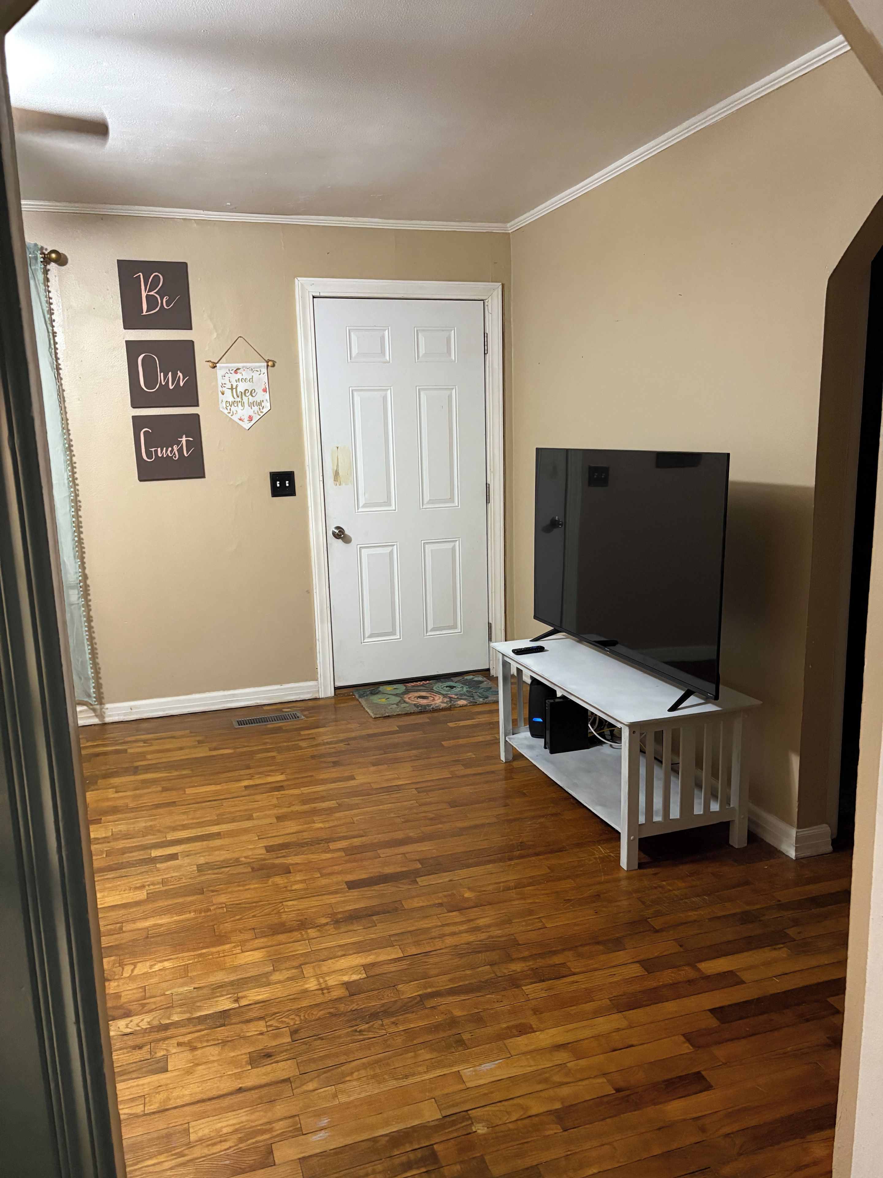 A room with a wooden floor, a white door, a flat-screen TV on a white stand, and a wall with a sign that reads 'Be Our Guest.'
