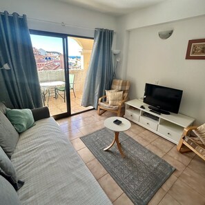 Ein Wohnzimmer mit einer Couch, einem Fernseher, einem kleinen Tisch und einem Sessel. Durch eine Glastür bietet sich ein Blick ins Freie.