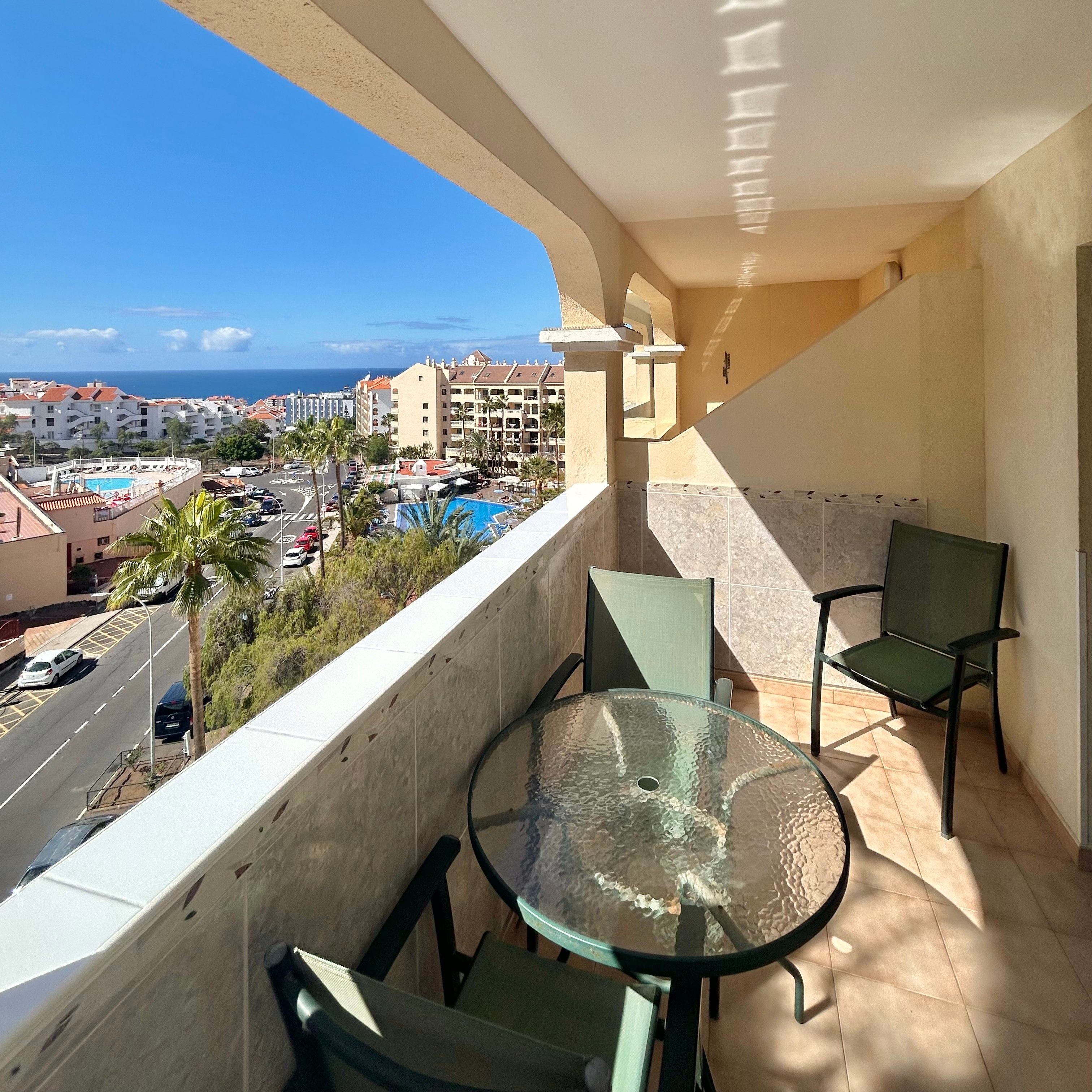 A balcony with a glass table and chairs, overlooking a pool and a cityscape.