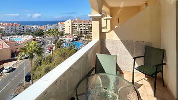 A balcony with a glass table and chairs, overlooking a pool and a cityscape.