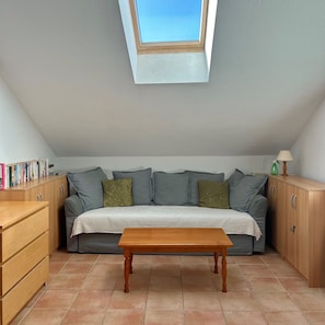 A cozy room with a sloped ceiling, a sofa with cushions, a wooden table, and a bookshelf.