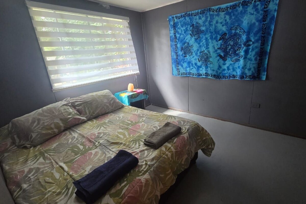 A bedroom with a bed, a nightstand, a window with blinds, and a blue tapestry on the wall.